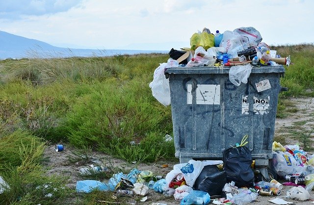 overflowing bin of trash in a empty field on a sunny day with a few clouds in the sky